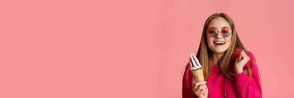 Smiling teenage girl in round glasses with ice cream in her hands gesturing and grimacing on pink studio background, panorama with copy space photo