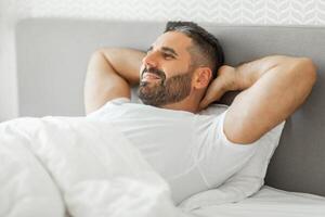 A man with a beard lies in bed with his hands behind his head, looking off to the side with a relaxed expression on his face. He is wearing a white t-shirt and is covered by a white duvet. photo