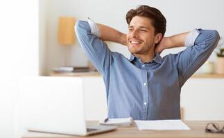 A smiling man sits at a desk with a laptop in front of him, relaxing with his arms behind his head. photo