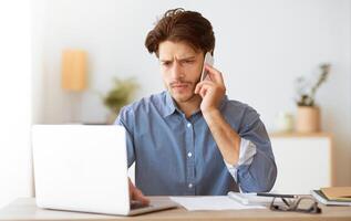 A man sits at a desk talking on his phone while working on his laptop. He looks focused and serious. photo