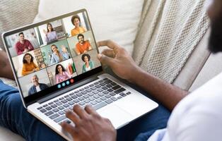 Black man sits on a couch, using a laptop to participate in a conference call with multiple other people. The man hands are visible as he engage with the screen. photo