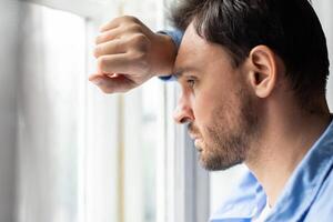 A man rests his forehead on his hand while looking thoughtfully out of a bright window, his expression conveys contemplation or possibly concern, closeup photo