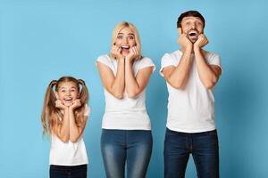 A young girl, her mother, and her father stand in front of a solid blue background. The family is smiling and looking up at the camera with their hands on their cheeks. photo