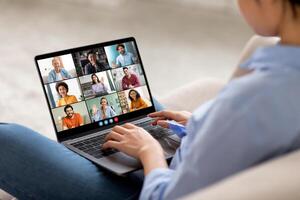 The image shows a woman sitting on a couch, participating in a conference on a laptop computer. Her laptop is open and shows a grid of nine feeds, each featuring a different person. photo