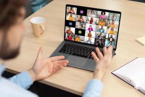 A man sits at a desk in front of a laptop, actively participating in a conference meeting with colleagues. His hands are raised, gesturing and engaging with the online participants. photo