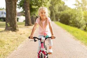 A young girl with blonde hair rides a pink bicycle on a paved path in a park. She is wearing a pink shirt and light-colored pants. The path is surrounded by green grass and trees photo
