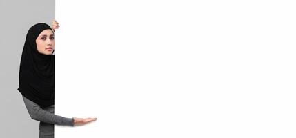 Serious arabic girl in headscarf pointing at white blank advertisement board with open palm, grey studio background photo