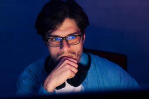 A young man wearing glasses sits in front of a computer in a dark room, thoughtfully resting his chin on his hand, likely deep in concentration on a project or task. photo