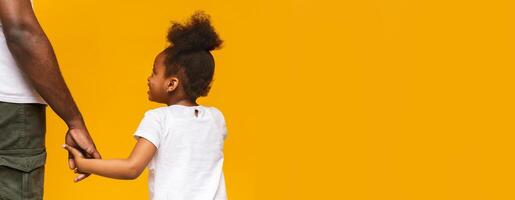 A joyful black child holds hands with a parent while walking together. The vibrant yellow backdrop enhances their connection, symbolizing warmth and love, copy space photo