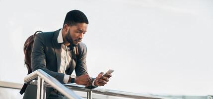 Black guy entrepreneur in a suit checking his phone while standing with a rolling suitcase outside a modern building, panorama with copy space photo