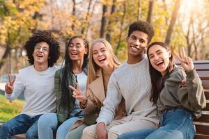 Positive international teenagers enjoying weekend at public park, sitting on bench and posing at camera photo
