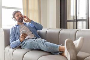 A man sits on a gray sofa, wearing headphones and a blue button-down shirt, while holding a smartphone in his hand. He has his legs propped up on the sofa and is smiling while listening to music. photo