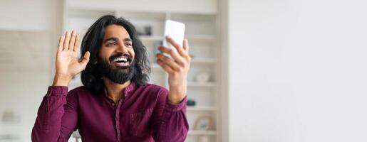 Smiling Indian freelancer man waving hand while having call on his smartphone, happy handsome eastern guy sitting at desk in modern home office, enjoying online communication, copy space photo