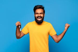 Hindu man with a beard is smiling and holding up a car key in his right hand while his left fist is clenched in the air. He is wearing a yellow t-shirt and is standing in front of a blue background. photo