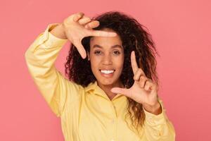 A young black woman with curly hair is smiling at the camera while making a framing gesture with her hands in front of her face, the background is a solid pink. photo