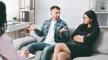 This image shows a couple in the middle of a heated argument during a therapy session. The man is gesturing wildly with his hands, while the woman has her arms crossed and looks at him photo