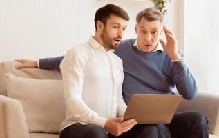Oops, computer problem. Shocked elderly man and his adult son using laptop looking at error message sitting on sofa indoor photo