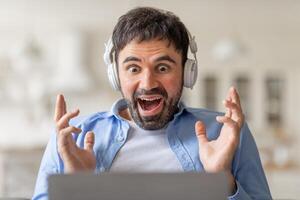 A man wearing headphones sits in front of a laptop with a wide open mouth and his hands raised in the air, showing excitement. He is looking at screen and appears to be engaged in something exciting. photo