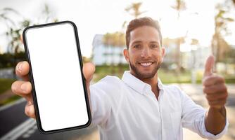 A man is standing outdoors, holding a smartphone with a blank screen, and giving a thumbs-up. He is smiling and looking directly at the camera. photo
