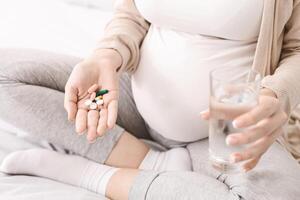 A pregnant woman is seen holding a pill in one hand and a glass of water in the other, preparing to take medication. The image shows the importance of taking prescribed medication during pregnancy photo