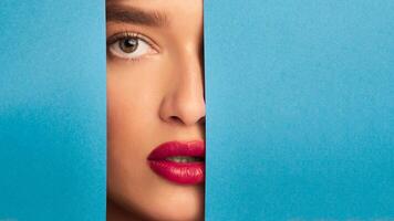 A woman face is partially visible as she peeks out from behind a piece of vibrant blue paper. Her eyes are wide open, looking directly at the viewer with a sense of curiosity and mystery. photo