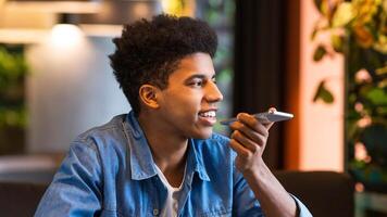 A young black guy is sitting in a modern cafe, holding his smartphone and using a voice assistant to perform tasks or send messages. photo