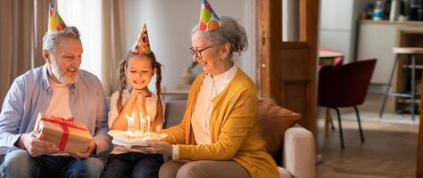 Three family members gather in a cozy living room to celebrate a birthday. The girl lights candles on a cake while the grandparents look on with joy, creating a heartwarming moment. photo