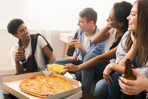 A group of diverse friends sitting in a circle around a large box of pizza, engaged in conversation and reaching out to grab a slice. The pizza box is open, revealing a variety of toppings. photo