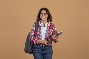 A young middle eastern woman wearing a red plaid shirt and blue jeans stands against a brown background. She is smiling at the camera, holding a stack of books in her right hand. photo