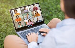 A person sits outside on a grassy lawn, using a laptop for a conference. The screen displays a grid of nine people, each in a separate frame photo