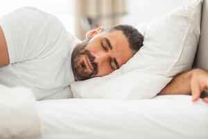 A man with a beard sleeps peacefully on his side, his head resting on a white pillow and his arm outstretched. The bedding is all white and the room appears to be a bedroom. photo