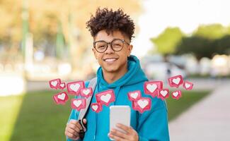 A young man with curly hair and glasses is smiling while holding a smartphone in his hand, and looking at the camera. He is standing outside during the daytime, and there are several red heart icons photo