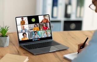 A person sits at a desk in a home office and attends a conference meeting on their laptop computer. The laptop screen displays a grid of tiles, showing the faces of multiple colleagues photo