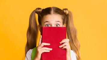 Back To School Anxiety. Scared Elementary Student Girl Hiding Behind Book On Yellow Studio Background. Free Space photo