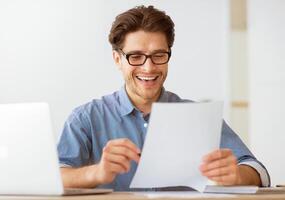 A man wearing glasses smiles and laughs while reading a document at his desk. photo