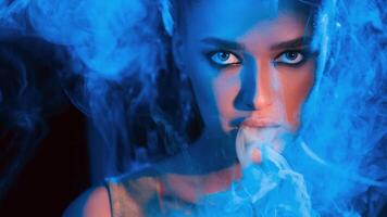 A woman with striking eyes is enveloped in blue smoke while she gazes intensely in a dark room. The blue lighting enhances the dramatic and mysterious atmosphere of the setting. photo