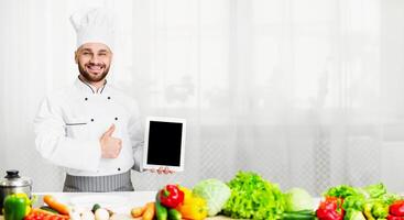 Great Cooking App. Chef Man Showing Tablet With Empty Screen Gesturing Thumbs Up Standing In Kitchen Indoor. Mockup photo