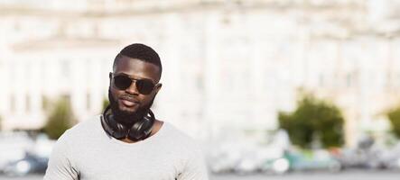 A man with a beard and sunglasses is wearing headphones around his neck. He is looking directly at the camera while standing in front of a blurred city background. photo