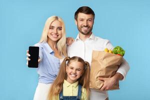 A happy family, consisting of a mother, father, and young daughter, stands together against a bright blue background. The mother holds a smartphone in front of her and smiles at the camera photo
