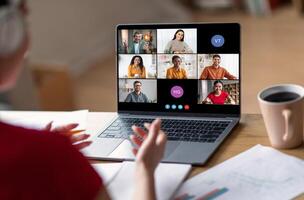A woman sits at her desk, participating in a conference call on her laptop. The screen displays multiple participants, with their names and initials visible photo
