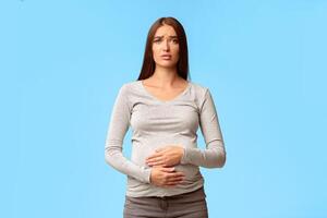 Pregnancy Stomachache. Sad Pregnant Woman Touching Belly Suffering From Pain Standing On Gray Background. Studio Shot photo