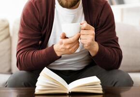 A man is seated on a couch, holding a cup in one hand and engrossed in reading a book. He appears relaxed and focused on the text, with a comfortable posture, cropped photo