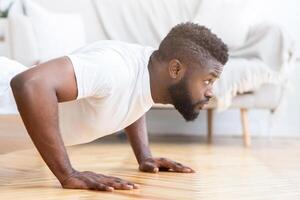 African American man in a white t-shirt is shown in profile performing a push up on a hardwood floor in his home. He is focused on the exercise and appears to be strong and determined. photo