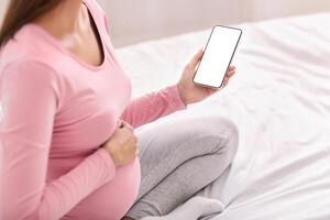 Pregnancy Applications. Unrecognizable Pregnant Lady Holding Cellphone With Empty Screen Sitting On Bed Indoor. Mockup, Cropped photo