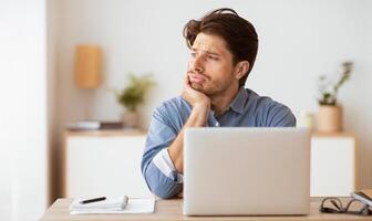 A pensive young man gazes out the window at his desk, chin on hand, with a laptop in front of him and a notepad and pen nearby. photo