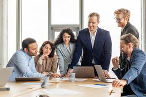 A group of diverse business professionals are gathered around a table in a modern office, working together on a project. They are looking at a laptop screen and discussing ideas photo