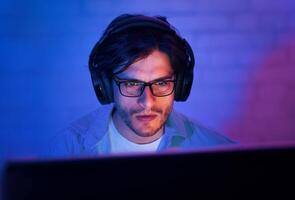 A young man wearing glasses and headphones sits in front of a computer screen in a dimly lit room. He appears to be playing a game, looking intently at the screen. photo
