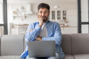 Man sitting on a couch in a home setting. He is looking at his laptop with a worried expression on his face and has his hands clasped together in front of him. He appears to be stressed or anxious. photo