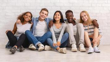 Multiracial happy students posing over white brick wall, sitting on floor and embracing, friendship concept photo