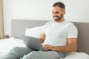 A man with a beard is sitting on a bed and using a laptop. He is wearing grey sweatpants and a white t-shirt. He is smiling and looking at the laptop screen. The bed is made with white sheets photo